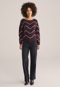 Bordeauxfarbenes Sweatshirt mit einem weißen Zickzackmuster, lockere Passform, lange Ärmel. Kombiniert mit schwarzen weit geschnittenen Jeans und Stiefeln mit Absatz.