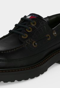 Μαύρο δερμάτινο boat shoe με κορδόνια, υφασμάτινες πλευρικές επιφάνειες, μεταλλικά ρουχισμένα, χοντρή σόλα και μικρό κόκκινο, λευκό και μπλε λογότυπο στη γλώσσα.