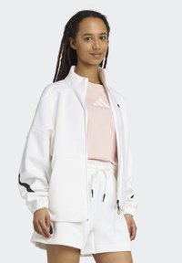 White zip-up jacket with black accents, worn over a light pink T-shirt and white shorts. Soft texture with elastic cuffs and pockets.