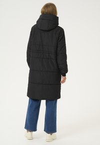 Manteau noir matelassé avec un col haut, des manches longues et des détails de coutures horizontales ; associé à un jean bleu à jambes larges et des baskets légères.