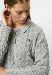 Žena s jemnými piehami, oblečená v svetlosivej svetríku pletenom vrkočovým vzorom s golierom a malým vyšívaným logom hráča polo.