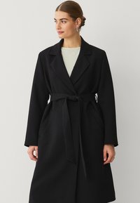 Sort lang frakke med bindebånd, brede revers og sidelommer, lavet af et blødt, struktureret stof. Simpelt, elegant design.