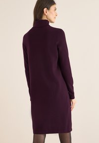 Vestido de cuello alto púrpura con mangas largas, diseño a la altura de las rodillas, textura suave y acentos minimalistas. Fondo liso para resaltar la prenda.