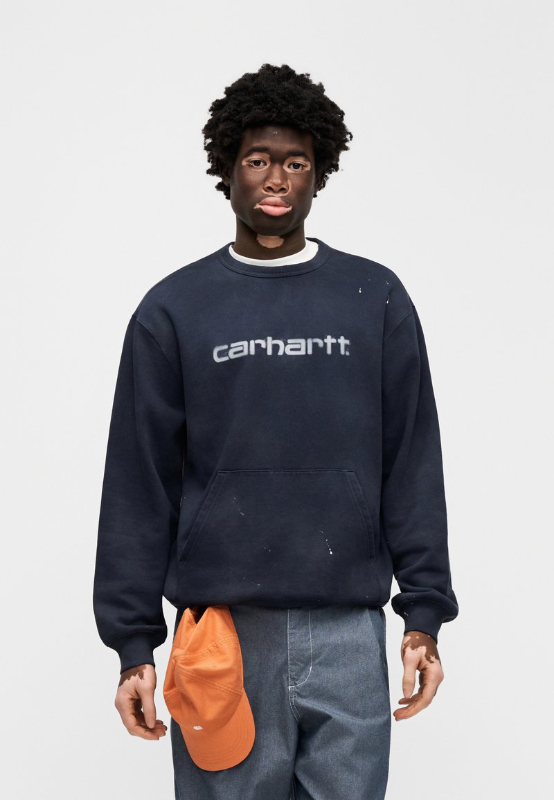 Marineblauer Sweatshirt mit "carhartt"-Logo, oversized Passform, mit einer Vorderfronttasche. Getragene blaue Jeans, orangefarbene Baseballkappe in die Hose gesteckt.