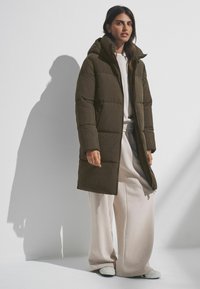 Manteau doudoune long à capuche en vert olive foncé, avec des lignes matelassées, une fermeture à zip et des poches latérales ; associé à un pantalon large de couleur claire.