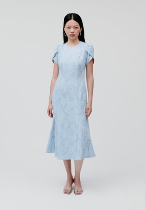 Lichtblauwe jurk met een bloemrijk textuurpatroon, korte puffmouwen en een uitlopende midi-lengte rok. Gecombineerd met doorzichtige haksandalen.
