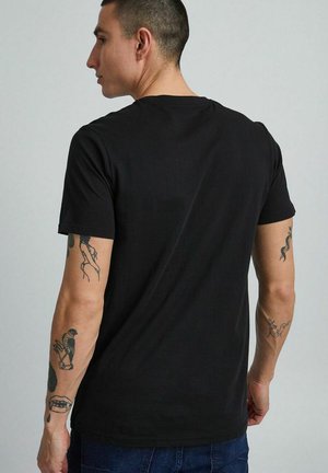 Mann mit kurzen Haaren, der ein schwarzes T-Shirt und blaue Jeans trägt, zeigt Tätowierungen auf beiden Armen, blickt nach links vor grauem Hintergrund.