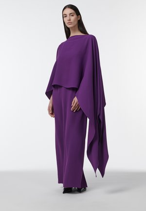 Mujer con cabello largo y oscuro, vistiendo una blusa púrpura fluida con mangas extendidas y pantalones anchos a juego, parada sobre un fondo blanco.