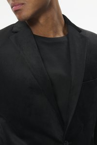 Mustast sametist bleiser nurgalöömise ja ühe nööbi sulguri, paaritatud musta t-särgiga all, mis näitab sujuvat tekstuuri.