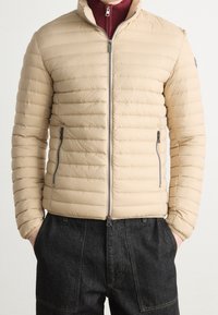 Beige pufferjacka med dragkedja fram, horisontell kviltad design, hög krage och två sidofickor. Buren över en vinröd tröja.