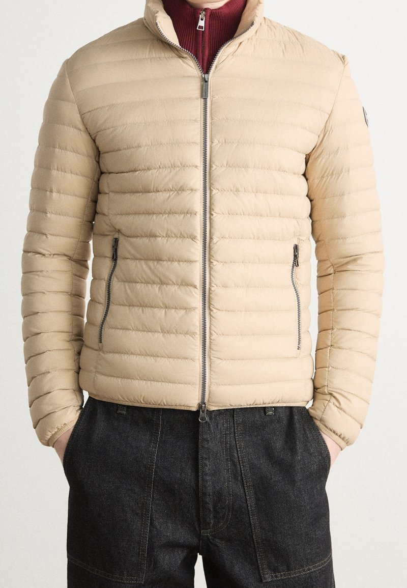 Beige pufferjacka med dragkedja fram, horisontell kviltad design, hög krage och två sidofickor. Buren över en vinröd tröja.