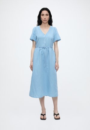 Donna in piedi che indossa un vestito midi azzurro chiaro a maniche corte con bottoni frontali, righe verticali bianche e sandali neri su uno sfondo semplice.