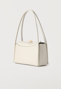 Kremasta pravokotna torbica z dolgimi zaobljenimi ročaji in zlato kroglo kot zaponko na enobarvnem ozadju.