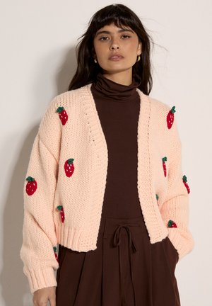 Mujer con cárdigan de punto grueso color rosa claro con patrones de fresas rojas sobre un suéter de cuello alto marrón oscuro y falda.