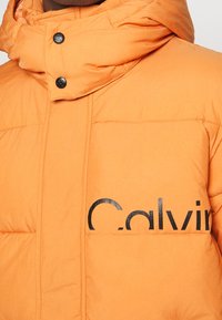 Pomarańczowa pikowana kurtka puchowa z wysokim kołnierzem, czarnymi zatrzaskami i wyrazistym czarnym logo na przodzie. Gładka tekstura i pikowane panele.