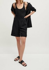 Zwart tanktopje, oversized zwarte blazer en op maat gemaakte zwarte shorts met een riem. Platte zwarte sandalen. Alle items hebben een effen kleur en een minimalistisch ontwerp.