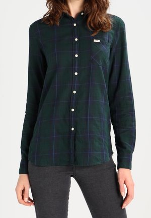 Ciemnozielona koszula w kratę z zapięciem na guziki, długimi rękawami, kieszenią na piersi oraz kontrastowymi białymi guzikami. Gładka tkanina o klasycznym kroju.