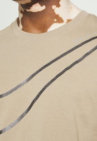 Krátký rukáv béžové tričko z bavlny s černými a kovově stříbrnými zakřivenými liniemi; text "just do it" ve stříbrné na rameni.