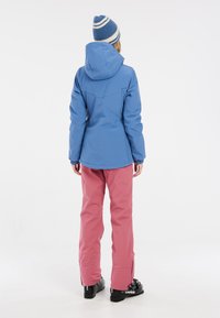 Blaue wasserfeste Jacke mit verstellbarem Kapuze, kombiniert mit pinken Skihosen mit Reißverschlüssen an den Knöcheln und schwarzen Skischuhen.