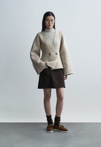 Bež jopič iz volnene mešanice z velikimi rokavi in dvema zlatima gumboma, nošen preko črtastega puloverja z visokim ovratnikom, kombiniran s rjavim semiš krilom.