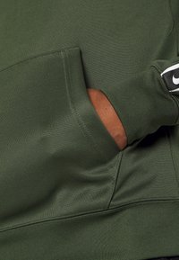 Felpa atletica verde con tasca frontale, dotata di polsini a costine e una patch nera del logo Nike sulla manica.