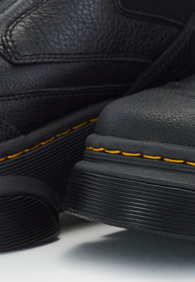 Scarpe in pelle nera con superfici testurizzate, dotate di una spessa suola in gomma nera e di un distintivo dettaglio di cucitura gialla lungo i bordi.