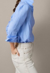 Chemise en lin bleu clair à manches longues avec poignets retroussés, associée à un pantalon de couleur crème doté de poches latérales et d'accents métalliques.
