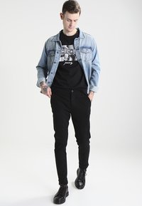 Light blue denim jacket over a black graphic t-shirt, paired with black tapered pants and black lace-up shoes. Featuring tattoos on the arm.