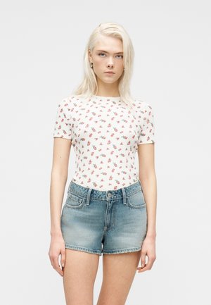 Blonde Frau trägt ein weißes kurzärmeliges Oberteil mit kleinem roten Blumenmuster und hellblaue hoch taillierte Jeans-Shorts.