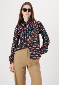 Svart bluse med et fargerikt biltrykk, med nakke-sløyfe, lange ermer og en avslappet passform. Kombinert med beige bukser.