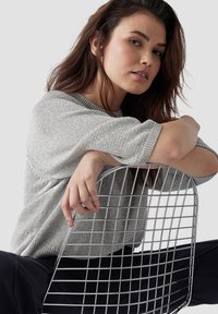 Grau gestrickter Pullover mit kurzen Ärmeln, kombiniert mit einem Metallgitterstuhl. Die Textur ist weich, und der Stuhl hat ein gitterartiges Design.