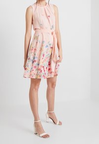 Hellrosa ärmelloses Kleid mit taillierter Passform, Floral-Muster und einem Gürtel in der Taille. Weiße Riemchen-High Heels runden das Outfit ab.