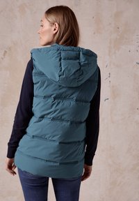 Teal pufferväst med huva, med horisontella sömmar och en mjuk textur. Matchad med en marinblå långärmad topp och jeans.