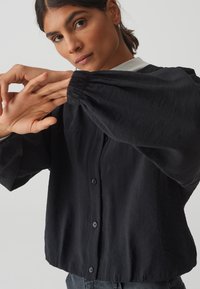 Zwarte button-up blouse met ballonmouwen, gemaakt van textuurstof. Kenmerkt zich door een afgeronde zoom en twee voorzakken.
