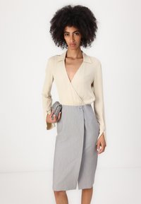 Blusa avvolgente beige con scollatura a V profonda, abbinata a una gonna grigia a lunghezza ginocchio con bottone laterale. Il modello tiene una borsa a mano argentata con texture.