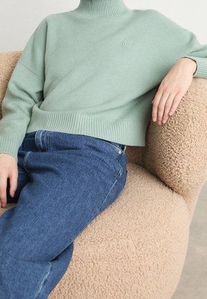 Pull turtleneck en tricot vert clair avec des poignets et un ourlet côtelés, associé à un jean en denim bleu, assis sur un fauteuil beige texturé.