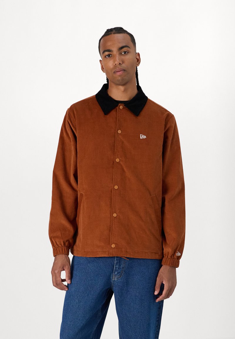 Brown corduroy jacket with a black collar, snap buttons, side pockets, and elastic cuffs. Small logo detail on the left chest.