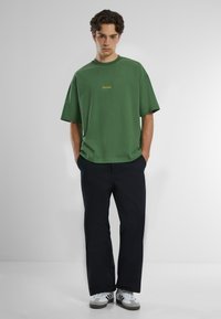 Zöld crew neck póló, laza szabással, sárga hímzéssel a mellkason. Sötét nadrággal és szürke sportcipővel viselve, fehér kiegészítőkkel.