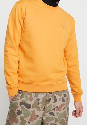 Oranger Crewneck-Sweatshirt mit gerippten Bündchen und Saum; mit einem kleinen Logo auf der Brust. Kombiniert mit Camouflage-musternden Hosen.