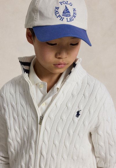 Junge trägt einen weißen, kabelgestrickten Zip-Pullover und eine weiße und blaue Polo Ralph Lauren Kappe, schaut mit neutralem Gesichtsausdruck nach unten.