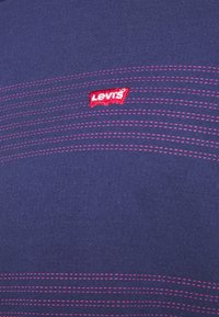 Gros plan d'un tissu bleu foncé avec des lignes roses en pointillés horizontaux et un petit patch rouge avec le logo Levi's au centre.