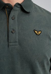 Grøn polo-shirt med et struktureret mønster, der har en knapkrave og et gulligt vinge-logo på venstre bryst.