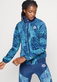 Navy en tealkleurig sportjack met een hoog kraag, ritssluiting en contrasterende texturen. Bevat een logo op de borst.