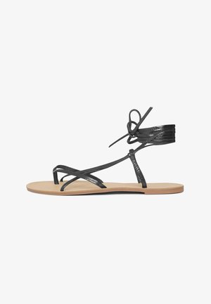 Sandales plates noires avec de fines lanières entrecroisées et une attache autour de la cheville. Texture lisse et brillante avec une semelle beige.