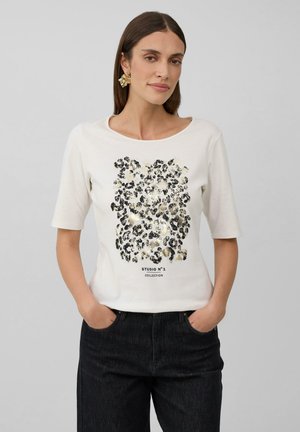 T-shirt blanc à manches courtes avec un design en imprimé léopard noir et or et le texte "COLLECTION STUDIO N°1" en bas. Texture : coton lisse.