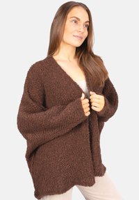 Femme aux longs cheveux bruns portant un cardigan oversized texturé brun sur un haut clair et un pantalon blanc, tenant les bords avant du cardigan.