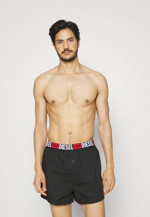 Boxer noir avec une ceinture de marque rouge et blanche, présentant une fermeture à bouton à l'avant, exposé sur un mannequin contre un fond uni.
