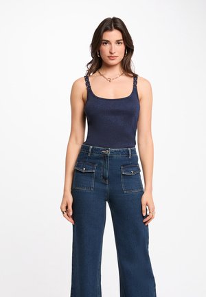 Giovane donna in piedi, indossa una canottiera blu navy senza maniche con spalline a catena e jeans a vita alta blu scuro con tasche frontali con patta.