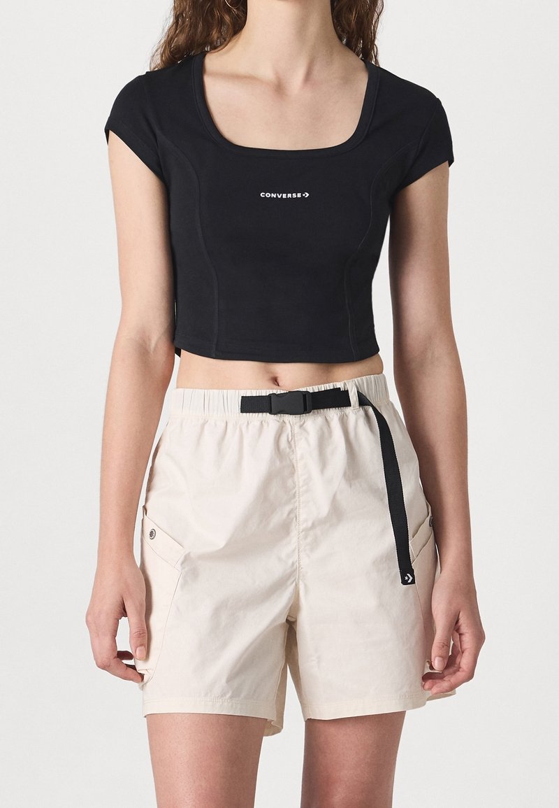 Personne portant un haut court noir à manches courtes avec le logo "CONVERSE" et un short beige taille haute avec ceinture noire ajustable.