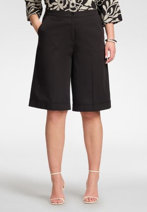 Mujer con pantalones cortos anchos negros hasta la rodilla con dobladillos puños y tacones beige con tiras, manos en los bolsillos y una blusa estampada.